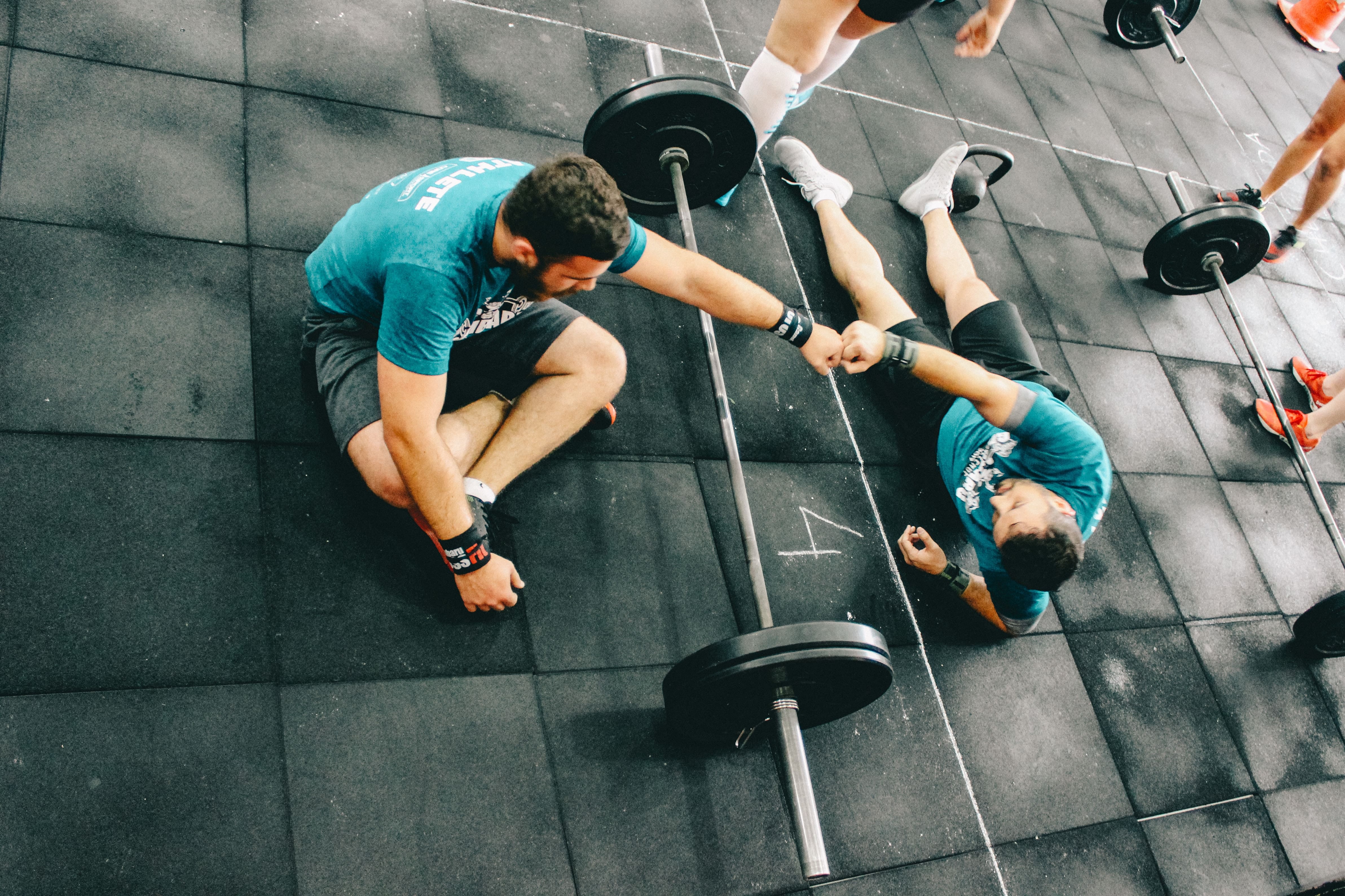Two men sitting on the floor with a barbell bar between them. They are fistbumping